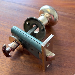 May include: A brass door knob set featuring a round handle, a dark green rectangular latch mechanism, and a metallic faceplate. The assembly includes a spindle and a small T-shaped thumb turn, showcasing a combination of polished brass and green finish.