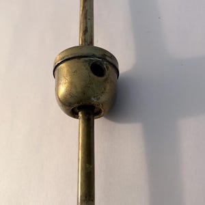 May include: A vintage brass object with a cylindrical body and a rounded top. The object has a long, slender stem extending from the top and bottom. The brass has a tarnished appearance, suggesting age. The object is likely a part of a larger mechanism.
