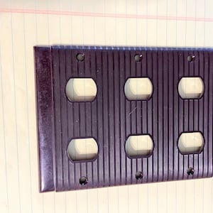 May include: A dark brown, rectangular electrical outlet cover with six outlet openings. The cover has a ridged texture and is designed to accommodate three dual electrical outlets. The cover is made of a durable material.