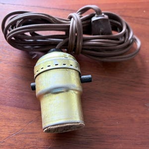 May include: A vintage brass-colored light socket with a brown electrical cord. The cord is coiled and has a standard plug. The light socket has two black side screws and a cardboard base. The items are on a wooden surface.