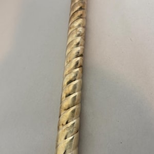 May include: A cylindrical metal tube with a polished gold-tone finish and a continuous diagonal twisted rope pattern running along its entire length. The object is straight, elongated, and features a hollow opening at the bottom end.
