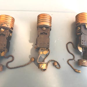 May include: Three vintage lamp socket replacements with pull chains. Each unit features a metallic threaded top, a dark rectangular body, and a brass-toned ball-end pull chain mechanism for manual operation, designed for electrical lighting fixtures.