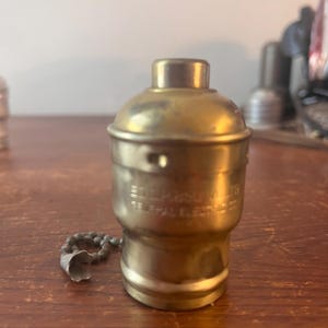 May include: A vintage brass light socket with a pull chain. The socket has visible text that reads "GENERAL ELECTRIC CO." and "SOL. M. 650 WATTS". The socket is cylindrical with a rounded top and a small chain.