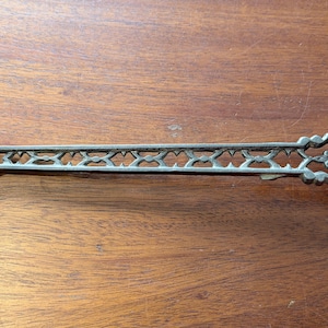 May include: A silver-toned metal tieback hook featuring an elongated, openwork lattice design along the arm, a curved hook at one end, and a threaded screw post at the opposite end for mounting into a wall or window frame surface.