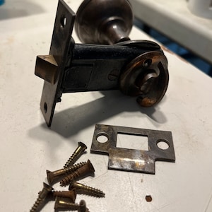 May include: A vintage door knob and strike plate with screws. The knob is made of dark metal and has a round shape. The strike plate is made of brass and has a rectangular shape. The screws are made of brass and have a Phillips head.