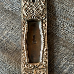 May include: Ornate, antique brass door pull with intricate floral and geometric designs. The rectangular plate features a keyhole and a recessed handle area. The metal has a tarnished, aged appearance, suggesting vintage craftsmanship.