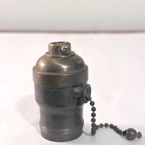May include: A metallic lamp socket with a dark, weathered finish featuring a pull chain mechanism. The word PRESS is embossed on the side near the pull chain entry point. The cylindrical component has a threaded top and a rounded base.