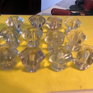 May include: A collection of clear, faceted glass cabinet knobs. Each knob has a diamond-shaped top and a cylindrical base. The knobs are arranged on a yellow surface, suggesting a hardware or home decor theme.