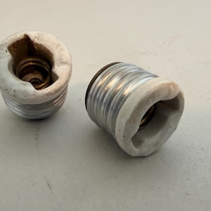 May include: Two light bulb socket adapters featuring a white ceramic exterior and a silver-colored metal threaded base. Each adapter has a hollow center with internal metal threading designed to hold a standard light bulb securely in place.