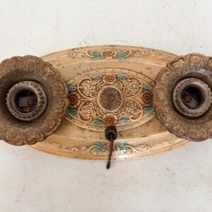 May include: An oval-shaped, beige ceiling light fixture base featuring two dark, textured, scalloped light sockets. The central plate is decorated with embossed floral patterns in muted orange and teal, and includes a small black pull chain.