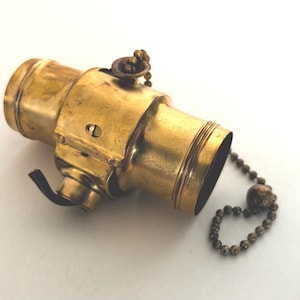 May include: A cylindrical brass lamp socket featuring a metallic finish with a visible patina. The component includes a small side-mounted switch mechanism and a beaded pull chain attached to the top, designed for controlling an electrical light bulb.