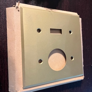 May include: A gold-colored metal wall plate with four holes and a rectangular cutout. The plate is packaged in a brown cardboard box.