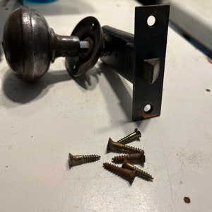 May include: An antique door knob and latch set with a dark, aged finish. The set includes a round knob, a circular plate, a rectangular latch plate, and several brass screws. The hardware is likely made of metal.