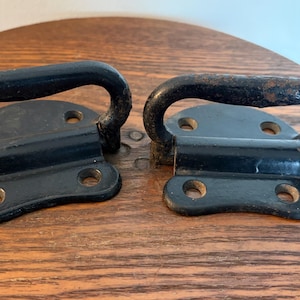 May include: Two black metal handles with a curved grip and a rectangular base with four screw holes. The handles are likely for a cabinet or drawer, and are set on a wooden surface.