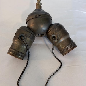 May include: Antique brass-toned three-way light fixture with three bulb sockets. Each socket has a pull chain with a decorative bell-shaped end. The fixture has a central ball-shaped component and a decorative top.
