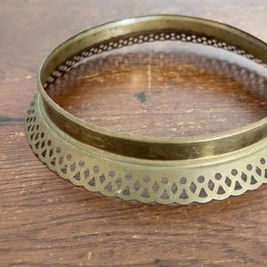 May include: A vintage brass decorative ring with a scalloped edge and openwork design. The ring is circular, with a slightly raised rim and a detailed pattern around the base. It sits on a wooden surface.