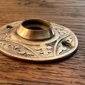 May include: A brass-colored, decorative metal object with a circular base and a raised central cylinder. The base features an embossed floral design. The object is likely a furniture component or decorative element.
