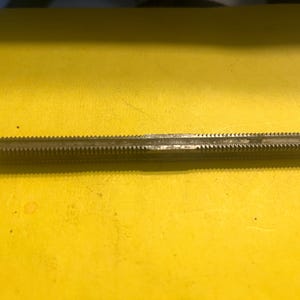 May include: A metal tap tool with a threaded shaft and sharp, angled teeth. The tool is a dark gray color and lies horizontally against a yellow background. The tap is used for cutting internal threads in materials.