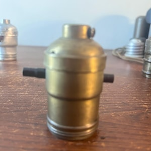 May include: A brass-colored light socket with a black switch. The socket has a cylindrical shape with a slightly wider base and a narrower top. Other light sockets are visible in the background.