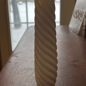 May include: A tall, off-white ceramic vase with a spiral design. The vase has a cylindrical shape and a glossy finish. The twisted pattern creates a textured surface. The vase is displayed on a wooden table.