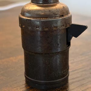 May include: A vintage, cylindrical light socket made of aged metal. The socket has a dark, rusty brown finish with a black lever on the side. The top is a lighter, tarnished color. The socket is designed to hold a light bulb.