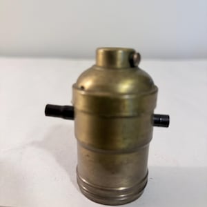 May include: A cylindrical brass light bulb socket featuring a metallic finish with a weathered patina. The component includes two small black plastic knobs extending from the sides and a threaded base for secure installation in a lamp fixture.