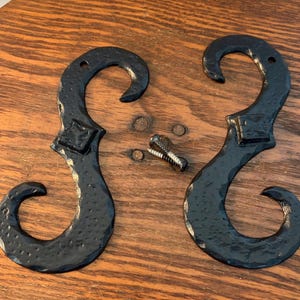 May include: Two black, wrought iron decorative hooks with a scroll design, diamond-shaped center, and mounting hardware. The hooks are displayed on a wooden surface.