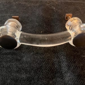 May include: Clear glass cabinet pull with a curved handle. The pull has rounded ends with dark circular accents and small square metal mounting plates.