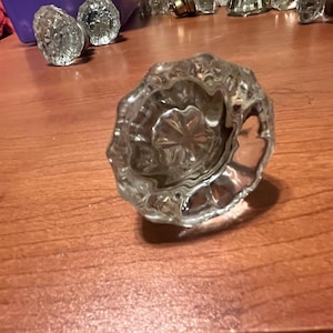 May include: Clear glass door knob with a flower-like design. The knob is faceted and reflects light, sitting on a wooden surface. Other similar knobs are visible in the background, suggesting a vintage or antique theme.