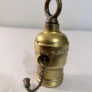 May include: A brass lamp socket featuring a pull chain with a small decorative end piece. The socket has a rounded top with a metal ring loop for hanging and a threaded base for a light bulb. The metal surface shows a metallic gold finish.