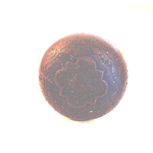 May include: A round, dark brown door knob featuring a central embossed quatrefoil pattern surrounded by intricate, swirling floral engravings. The surface has a weathered, textured finish with visible signs of age and wear across the circular face.