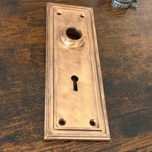 May include: A rectangular, antique-style door plate with a copper finish. The plate features a keyhole, a circular opening for a doorknob, and four screw holes. The edges are decorated with a detailed, raised pattern.