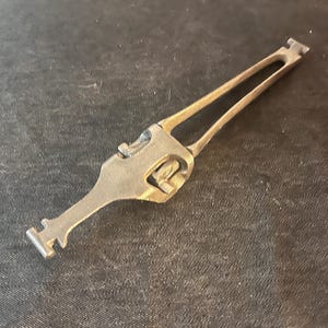 May include: A metallic, antique-looking tool with a unique shape. It features a long, slender handle that splits into two parallel arms. The tool has a complex head with a central opening and a small, rectangular end. The object is likely a specialized instrument.