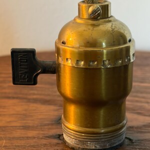 May include: A vintage brass lamp socket with a black switch labeled "Leviton." Two twisted wicks protrude from the top. The socket is set against a wooden surface, showcasing its aged patina and classic design.