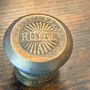 May include: Close-up of a vintage brass bottle stopper. The top is round with the word "ROYAL" in the center, surrounded by text and dates. The stopper has a mesh-like design on the lower portion.