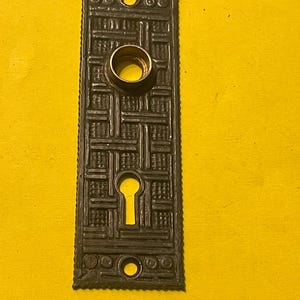 May include: A dark, rectangular antique door plate with an intricate geometric pattern. It features a keyhole, a circular opening, and two smaller holes. The plate has a bronze finish and is set against a yellow background.