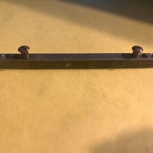 May include: A rectangular metal bar with two screws. The bar is dark with a slightly worn appearance, and the screws are a darker color. The bar has three holes on each end. The background is a solid yellow color.