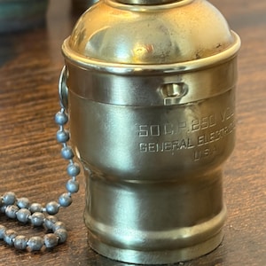 May include: A vintage brass lamp socket with a pull chain. The socket has the text "50 C.P. 250 V GENERAL ELECTRIC USA" engraved on its side. The metal has a tarnished, aged appearance.