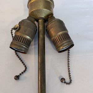 May include: A vintage brass lamp fixture with three light sockets. The fixture has a central pole with two sockets angled to the sides and one on top. Each socket has a pull chain with a small ball at the end.