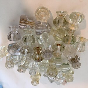 May include: A collection of clear glass cabinet knobs featuring a variety of shapes, including faceted, round, and hexagonal designs. Each knob has a central hole for mounting, with some showing subtle internal variations in the glass clarity.