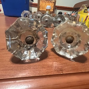 May include: Clear, faceted glass cabinet knobs with a central, dark-colored attachment. The knobs have a starburst design and are arranged on a wooden surface. Other knobs are visible in the background, suggesting a set.