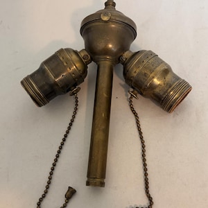 May include: A vintage brass lamp socket cluster featuring a central vertical stem topped with a finial, two side-facing light bulb sockets, and two hanging metal pull chains with small decorative ends, all displaying a tarnished metallic finish.