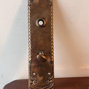 May include: Antique brass door plate with ornate detailing and a keyhole. The plate has a rope-like border and a decorative knob.