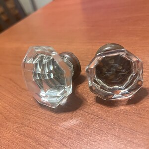 May include: Two clear, faceted glass door knobs with a vintage design. The knobs have a crystal-like appearance and are mounted on a dark metal base. They are placed on a wooden surface.