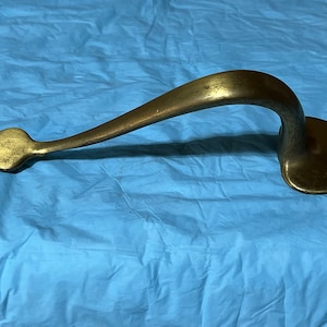 May include: A vintage-style brass door handle. The handle has a curved design with a pointed end and a heart-shaped base with a square hole. The brass has a slightly aged, tarnished appearance.