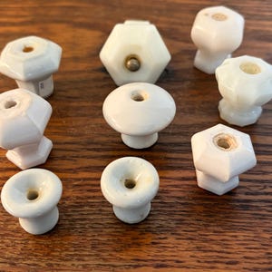 May include: A collection of white ceramic cabinet knobs in various shapes, including round, hexagonal, and faceted designs. Each knob has a small hole in the center and is set against a warm, wooden surface.