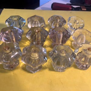 May include: A collection of twelve clear glass cabinet knobs with a faceted, flower-like design. The knobs have a vintage aesthetic and are arranged on a yellow surface, showcasing their intricate details and potential for home decor projects.