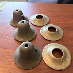May include: Six antique brass lamp parts, including three bell-shaped shades and three circular trim pieces. The shades have a dark, aged patina, while the trim pieces are a lighter brass color. The parts are arranged on a wooden surface.