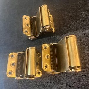 May include: Three brass-colored spring hinges are displayed against a dark blue background. Each hinge has a rectangular base with screw holes, a cylindrical barrel, and a spring mechanism. The hinges appear to be new and unused.