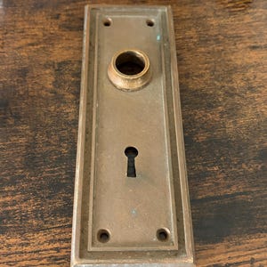 May include: A rectangular antique door plate with a keyhole and a circular opening for the doorknob. The plate is made of bronze-colored metal and has a rectangular shape with rounded corners. Four screw holes are visible.